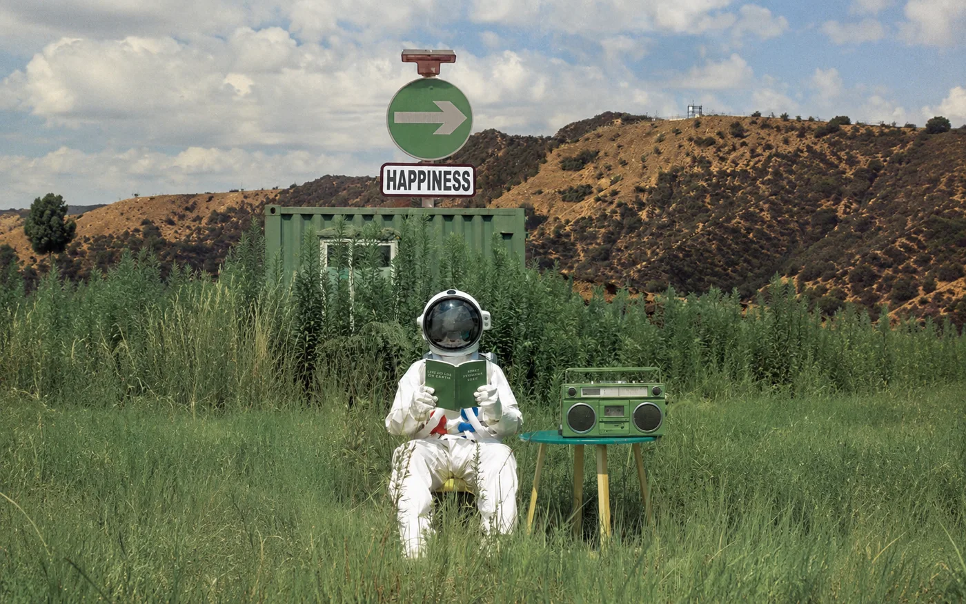 Astronauta sentado num campo verde lendo um livro, ao lado de uma placa que aponta para 'Happiness'